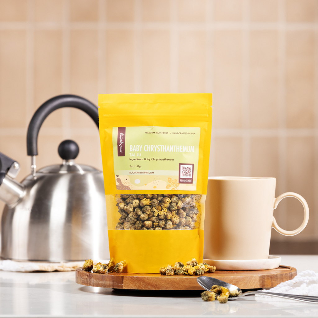 A lifestyle image of Baby Chrysanthemum herbal tea displayed on a wooden tray next to a stainless steel kettle, a beige ceramic mug, and loose dried flowers on a spoon.