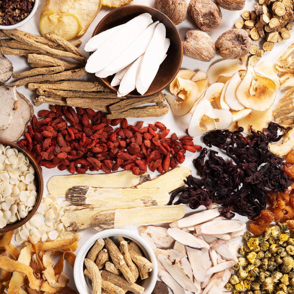 An overhead flat-lay arrangement of various dried and fresh herbal ingredients arranged around a white central background.